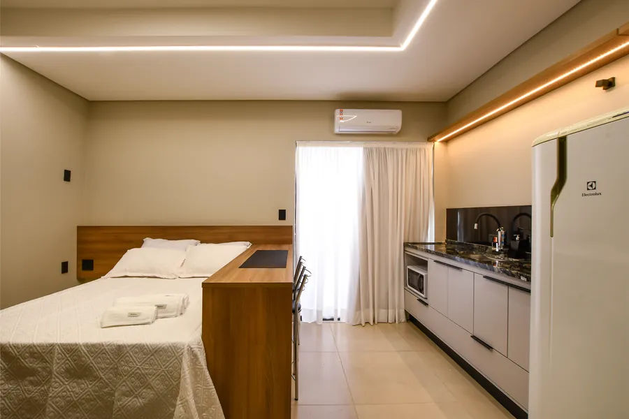 Cama com enxoval branco e cozinha equipada.
